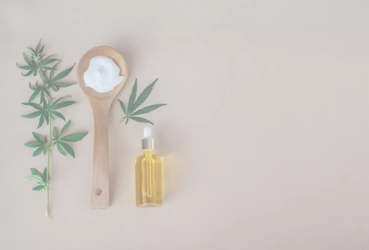 Hemp plant beside wooden spoon that has cream in it with an oil bottle beside the spoon to the right