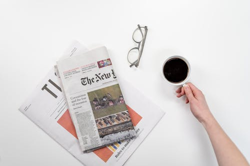 A female hand grabbing a coffee with a news paper beside the coffee and glasses in between both