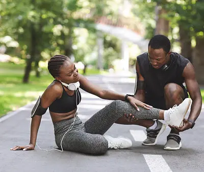 photo of a young man and woman stopped on a jogging path. the woman is in pain and the man is investigating her sprained or strained ankle.